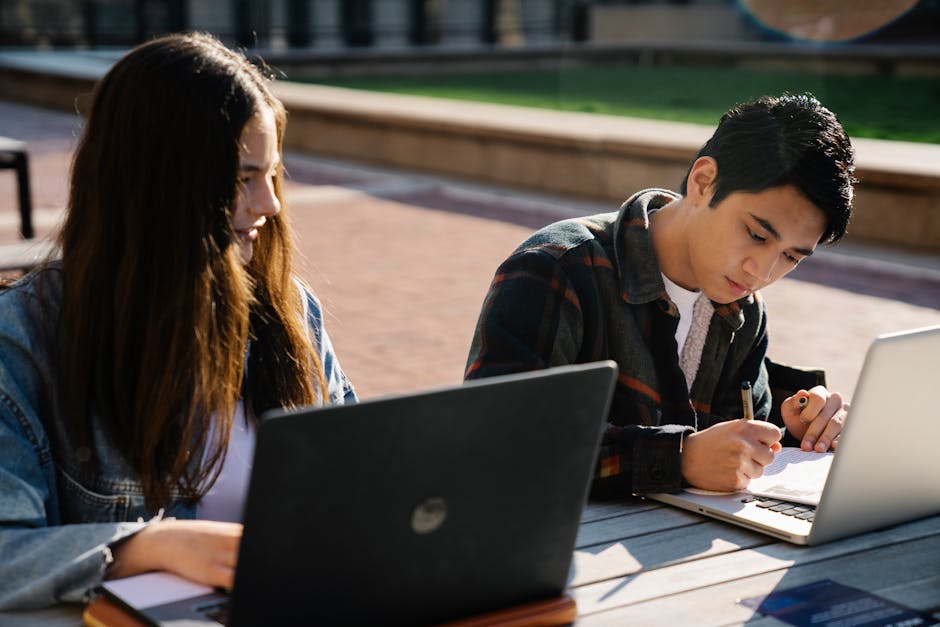 Two students engaged in study session outdoors, using laptops and writing notes